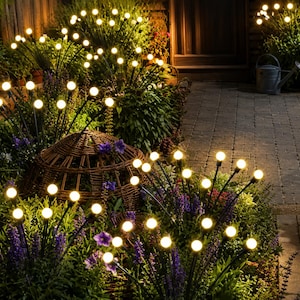 Pode incluir: Várias luzes de jardim solares com hastes pretas finas rematadas com lâmpadas esféricas de luz branca quente. Estas luzes estão dispostas em grupos por todo o jardim, entre folhagem verde, flores roxas e um cesto de vime.