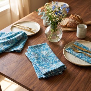May include: A set of blue and white floral patterned cloth napkins featuring a repeating botanical design. The napkins are displayed folded on a wooden table alongside ceramic plates, gold-toned cutlery, and a glass vase filled with flowers.