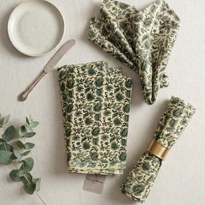 Puede incluir: Un juego de servilletas de tela color crema con un estampado floral en verde oscuro. El conjunto incluye servilletas dobladas y enrolladas, una de ellas sujeta con un servilletero de metal dorado, junto a un plato pequeño, un cuchillo y una rama.