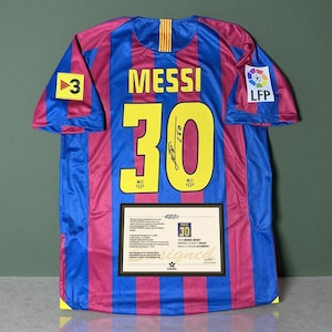 May include: A blue and red striped football shirt featuring the name MESSI and the number 30 in yellow on the back. The shirt includes a black signature on the number 30, a LFP patch, a 3 patch, and a rectangular certificate of authenticity.