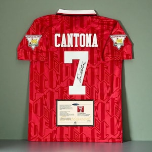 Puede incluir: Una camiseta de fútbol roja del Manchester United con cuello blanco, el nombre CANTONA y el número 7 en blanco. La camiseta incluye una firma negra, dos parches de Champions 1992-1993 en las mangas y un certificado de autenticidad rectangular.