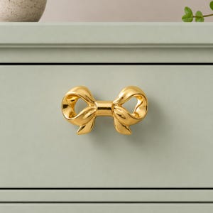 May include: A polished gold-toned metal drawer pull shaped like a decorative bow. The hardware features a smooth, reflective surface with curved loops and a central knot, mounted horizontally on a flat, light sage green cabinet drawer front.