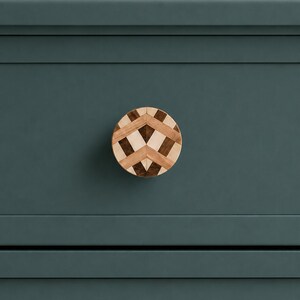 May include: A circular wooden cabinet knob featuring a geometric inlay pattern of light and dark wood segments. The knob is mounted on a flat, matte dark teal surface with horizontal recessed lines running across the panel.