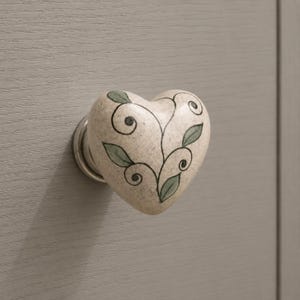 May include: A heart-shaped cabinet knob with a speckled beige finish, featuring a hand-painted design of black swirling vines and muted green leaves. The knob is attached to a circular metal base, providing a functional handle for furniture.