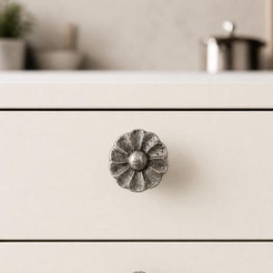 May include: A circular cabinet knob with a flower-shaped design and a textured, weathered silver-toned finish. The knob is mounted on a smooth, light-colored drawer front, featuring a central raised dome surrounded by petal-like segments.