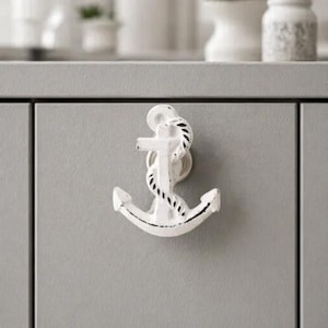 May include: A white cabinet knob shaped like a nautical anchor with a distressed finish. The anchor features a rope detail wrapped around the central post and is mounted onto a flat, light gray cabinet door surface with a vertical seam.