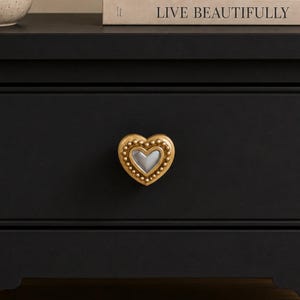 May include: A heart-shaped drawer knob featuring a metallic gold finish with a beaded border and a smooth, light-colored center inlay. The knob is mounted on a solid black drawer front, providing a high-contrast look for furniture hardware.