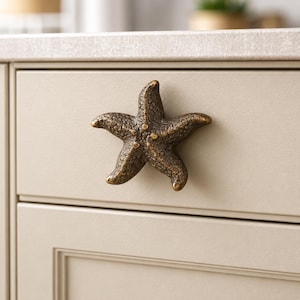 May include: A bronze-toned starfish-shaped cabinet knob with a textured, weathered surface. The five-pointed sea creature design is mounted onto a flat, light beige drawer front, featuring a dark metallic finish with subtle highlights.