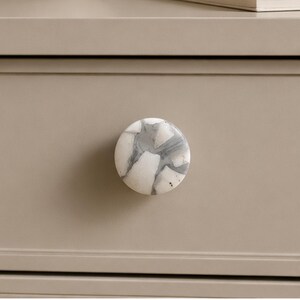 May include: A round cabinet knob featuring a mosaic-style pattern of white and grey stone-like segments. The knob has a smooth, polished surface and is mounted onto a flat, solid beige drawer front with horizontal trim detailing.