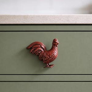 May include: A copper-toned metal cabinet knob shaped like a rooster in profile, featuring detailed plumage textures on the wings and body, mounted on a flat sage green drawer front with a horizontal panel line and a light-colored countertop edge.