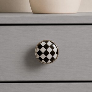 May include: A round cabinet knob featuring a black and white checkered pattern on its face. The circular knob has a smooth, glossy finish and is framed by a thin, metallic silver-toned border, mounted onto a flat, light gray wooden drawer front.