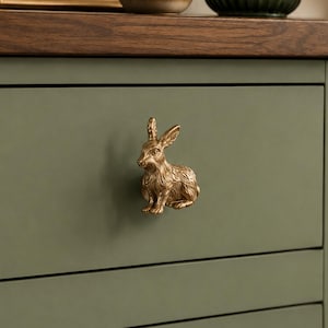 May include: A metallic gold-toned cabinet knob shaped like a sitting rabbit. The rabbit figure features detailed fur texture and pointed ears, mounted onto a matte olive green drawer front with a horizontal wood grain top edge.