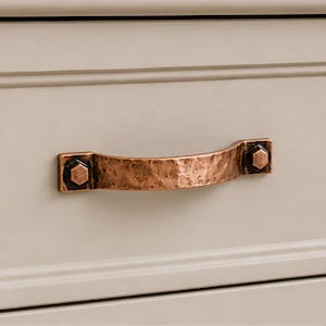 May include: A hammered copper drawer pull with a curved handle and two hexagonal bolt-style mounting points, attached to a light beige cabinet drawer. The metal finish features a textured, uneven surface with dark brown accents in the crevices.