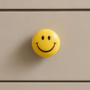 May include: A round, bright yellow cabinet knob featuring a black printed smiley face graphic with two oval eyes and a wide, curved mouth. The knob has a smooth, glossy finish and is mounted onto a flat, light beige surface with horizontal lines.