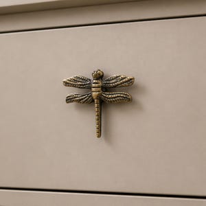 May include: A dragonfly-shaped drawer knob made of antique brass-toned metal. The knob features detailed wings with etched lines and a segmented body, mounted on a smooth, solid light beige drawer front surface.