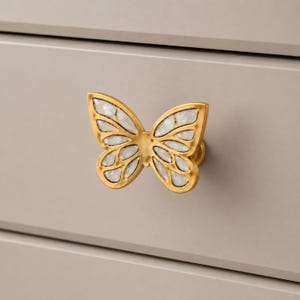 May include: A gold-toned butterfly-shaped drawer knob featuring iridescent white inlay segments within the wings. The hardware is mounted onto a smooth, light gray surface, showcasing a symmetrical design with a metallic finish.