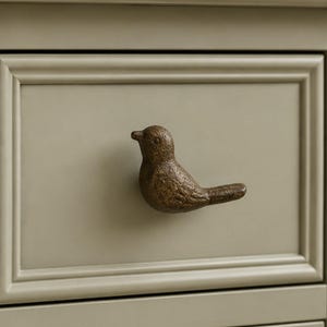 May include: A brown, bird-shaped drawer knob with a textured, weathered finish. The knob is mounted on a light beige, rectangular drawer front that features a raised, framed border design, providing a functional handle for the furniture piece.