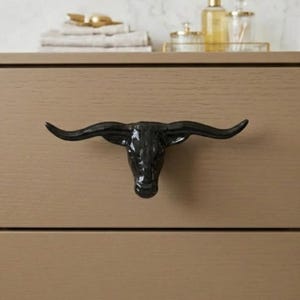 May include: A black cabinet pull shaped like a longhorn bull head with wide, curved horns. The hardware is mounted on a light brown wooden drawer front, featuring a glossy finish that reflects light across the surface of the animal head.