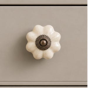 May include: A flower-shaped cabinet knob with a glossy off-white finish and subtle speckled texture. The center features a circular, dark bronze-toned metal plate with a ridged, sunburst-like pattern and a small, smooth, rounded metal knob.