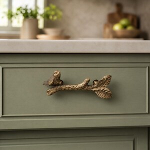 May include: A brass-toned metal cabinet pull shaped like a textured tree branch with a small bird perched on one end and detailed leaves on the other. The hardware is mounted horizontally on a flat, sage green cabinet drawer front.