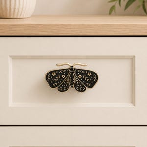 May include: A moth-shaped drawer knob featuring a matte black finish with gold-toned metal edges. The surface of the moth is decorated with engraved white floral and geometric patterns, including small flowers, dots, and chevron-like lines.