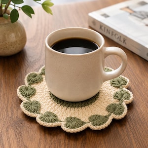 May include: A round, crocheted coaster featuring a cream-colored center and a scalloped border of dark green leaf-shaped motifs. A speckled, off-white ceramic mug filled with black coffee sits centered on top of the textured, handmade coaster.