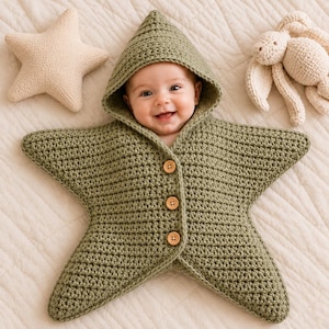 May include: A sage green crocheted star-shaped baby bunting with a hood and three light brown wooden buttons down the front. The garment features a textured knit pattern and is shaped like a five-pointed star, providing full coverage for the infant.
