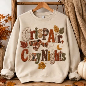 Peut inclure: Un sweat-shirt à col rond couleur crème portant l'inscription "Crisp Air, Cozy Nights" dans une police de style patchwork aux textures variées. Le motif inclut des éléments automnaux comme des carreaux, des feuilles brodées et une écharpe.