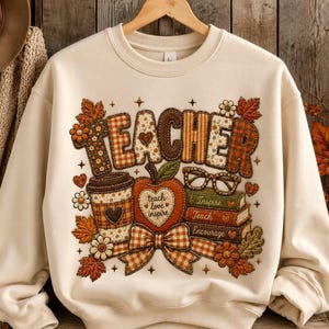 May include: A cream-colored crewneck sweatshirt featuring a graphic print with the word TEACHER in a patchwork style. The design includes a coffee cup, an apple with the text teach love inspire, a stack of books labeled Inspire, Teach, Encourage, and glasses.