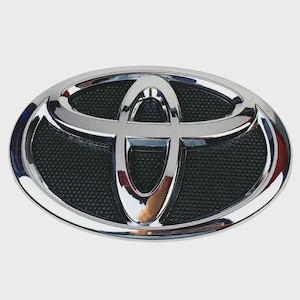 May include: An oval-shaped Toyota vehicle emblem featuring a polished chrome finish on the iconic three-ring logo. The background of the emblem consists of a textured black grid pattern, providing a high-contrast look to the metallic silver surface.