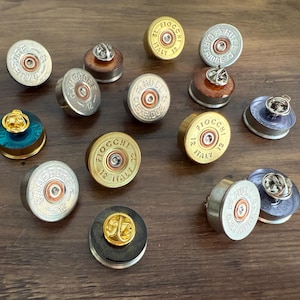 May include: A collection of twelve circular lapel pins crafted from repurposed 12-gauge shotgun shell bases. The pins feature metallic finishes in gold, silver, and blue, with embossed text including Fiocchi Italy and Hull Cartridge.