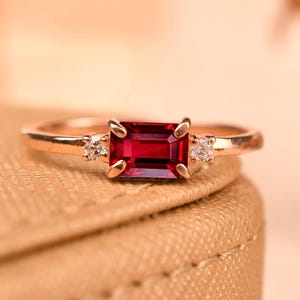 Könnte beinhalten: Ein Ring aus Roségold mit einem zentralen, rechteckigen, im Smaragdschliff geschliffenen roten Edelstein, der von vier Krappen gehalten wird, flankiert von zwei kleinen runden, klaren Steinen auf jeder Seite der Ringschiene.
