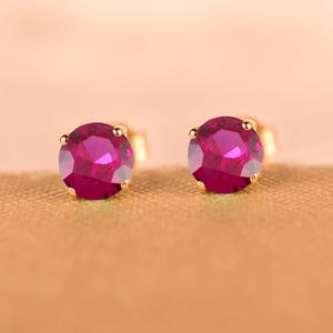 Könnte beinhalten: Ein Paar runde Ohrstecker mit leuchtend magentafarbenen Steinen, die in goldfarbenen Metallfassungen sitzen. Jeder Ohrring besteht aus einem runden, facettierten Edelstein, der sicher von vier kleinen Metallkrallen gehalten wird.