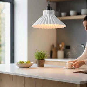 May include: A white pendant light with a pleated, cone-shaped shade and a black cord fixture. The lamp features a textured, speckled finish and hangs from a black ceiling mount, casting a warm glow downward from its wide, fluted opening.