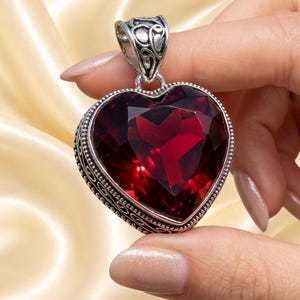 May include: A heart-shaped pendant featuring a deep red faceted gemstone set in a silver frame with intricate scrollwork detailing along the edges and a decorative bail at the top, showcasing a polished metallic finish and a vibrant crimson hue.
