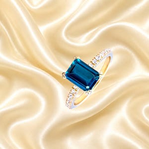 Può includere: Un anello in oro con una grande pietra preziosa rettangolare blu scuro a taglio smeraldo come pietra centrale. Il gambo è realizzato in oro ed è impreziosito da una fila di piccole pietre bianche rotonde e trasparenti.