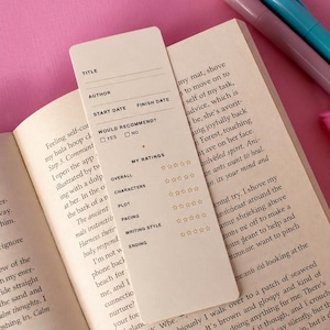 Puede incluir: Un marcapáginas de papel color crema con campos impresos para título, autor, fecha de inicio, fecha de finalización y una casilla de verificación de recomendación. Debajo, una sección de calificación presenta cinco categorías con iconos de estrellas vacíos para general, personajes, trama, ritmo, estilo de escritura y final.