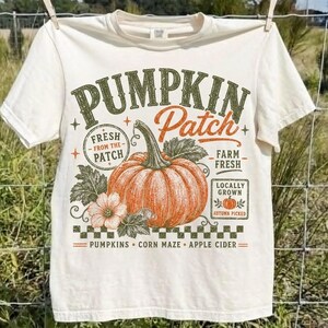 Op de afbeelding: Een crèmekleurig t-shirt met een grafische print met de tekst PUMPKIN Patch, FRESH FROM THE PATCH, FARM FRESH, LOCALLY GROWN, AUTUMN PICKED, PUMPKINS, CORN MAZE en APPLE CIDER, naast een illustratie van een grote oranje pompoen.