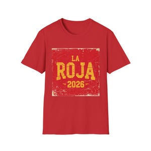Può includere: Una t-shirt rossa a maniche corte con una grafica quadrata sul petto. La grafica presenta uno sfondo rosso usurato con il testo giallo LA ROJA e 2026 stampato in un carattere grassetto e squadrato. Il design ha un aspetto invecchiato.