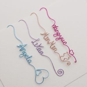 May include: Four custom wire bookmarks in blue, purple, copper, and pink. Each bookmark features a name written in cursive wire script: Angela, Lilaa, MinMin, and Mangyue. The wire ends are shaped into decorative hearts, swirls, and a cat outline.