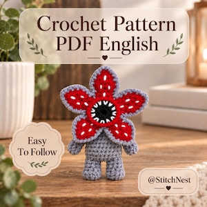 May include: A crochet pattern PDF in English featuring a small, handmade amigurumi creature with a grey body and a large, five-petaled red flower-like head. The head has a black center with white teeth-like details and white stitch accents.