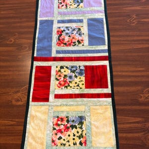 May include: A rectangular quilted table runner featuring four central square panels with a floral print of red, blue, and yellow flowers. The runner is pieced together with solid blocks of lavender, blue, red, and yellow fabric and light green borders.