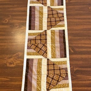 May include: A rectangular quilted table runner featuring a geometric pattern of alternating blocks. The design incorporates strips of fabric in shades of brown, tan, and cream, accented with plaid and floral prints, all bordered by a solid white trim.
