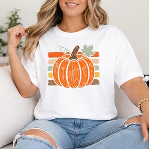 May include: A white short-sleeve t-shirt featuring a central graphic of a bright orange pumpkin with a brown stem and a light green leaf. The pumpkin is layered over horizontal stripes in shades of orange, yellow, and brown.