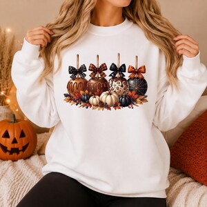 Op de afbeelding: Een wit sweatshirt met ronde hals en een grafische print van vier karamelappels met strikken, omringd door herfstbladeren, kleine pompoenen en bessen. De appels hebben diverse patronen, waaronder stippen, ruiten, luipaardprint en spinnenwebben.