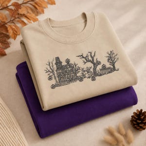 May include: A beige crewneck sweatshirt featuring a black embroidered illustration of a haunted house, bare trees, and pumpkins, layered on top of a solid purple sweatshirt. The design depicts a multi-story house with a fence and foliage.