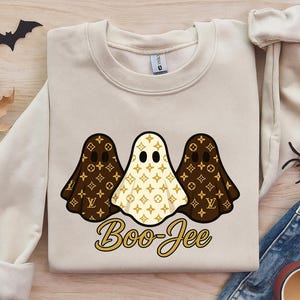 May include: A cream-colored crewneck sweatshirt featuring a graphic of three ghosts with a brown and cream repeating monogram pattern. The text "Boo-Jee" is printed in gold below the ghosts. The sweatshirt has long sleeves and a ribbed neckline.