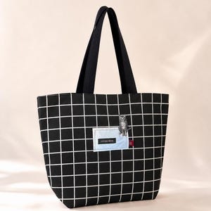 Puede incluir: Un bolso tote negro con un patrón de cuadrícula blanca, dos asas negras y un parche rectangular azul claro con el texto ESPANA ODARE. Una pequeña ilustración de un gato gris está situada en el borde superior del parche azul claro.