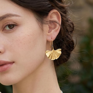 May include: A pair of gold-toned drop earrings shaped like ginkgo leaves. The earrings feature a textured, fan-like surface with delicate veining, suspended from thin, curved metal hooks that hang from the earlobe in a dangling style.