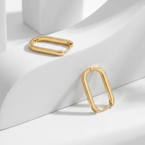 May include: A pair of gold-toned hoop earrings featuring a smooth, elongated rectangular shape with rounded corners. The earrings have a hinged closure mechanism and a polished metallic finish, presented as two separate pieces in a set.